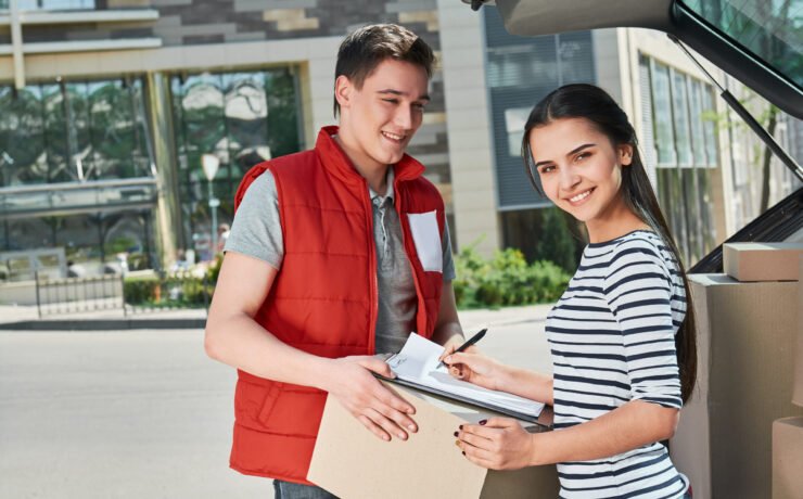 Cheerful man wearing red postal uniform is delivering parcel to a satisfied client. He brought her a parcel directly to the trunk of her car. Dark-haired woman is signing receipt of delivery package. Friendly worker, high quality delivery service. Outdoors.